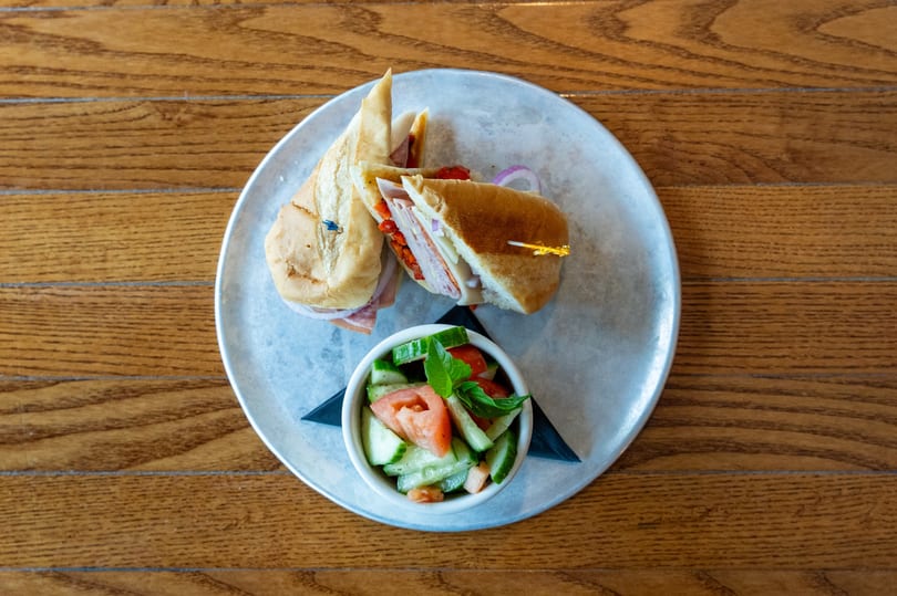 Two halves of a deli sandwich with a side of cucumber and tomato salad on a metal plate.