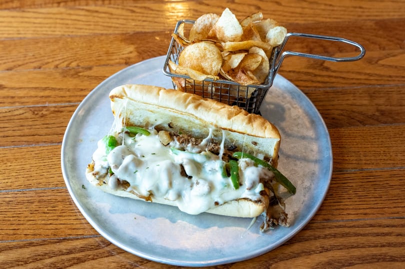 Cheesesteak sandwich with melted cheese, green peppers, and onions, served with a side of chips.