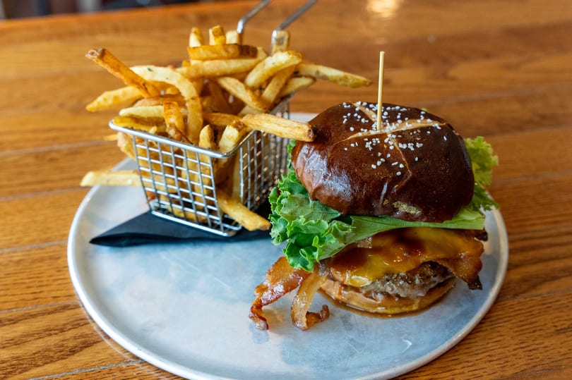 A gourmet burger with bacon, cheese, lettuce, and a pretzel bun, served with a side of fries.