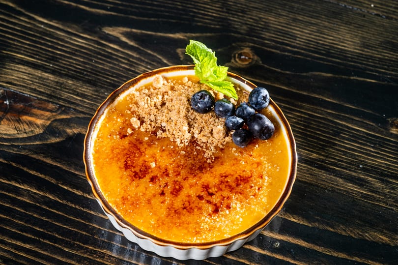 Crème brûlée dessert in a ramekin, topped with blueberries, mint, and crumble on a dark wooden table.